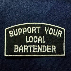 May include: A black patch with white embroidered text that reads "SUPPORT YOUR LOCAL BARTENDER". The patch has a white border and is set against a dark blue fabric background.