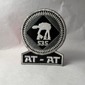 May include: A black and white embroidered patch featuring an AT-AT walker from Star Wars. The patch has a black diamond shape with a grey border and a white outline. The text "AT-AT" is on a black rectangle below the walker. The walker is numbered "535".