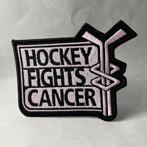 May include: Black and white embroidered patch with the text "Hockey Fights Cancer" in pink lettering. The patch features two crossed hockey sticks.