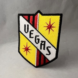 Può includere: Una toppa ricamata gialla e rossa con un bordo nero a forma di scudo. La toppa ha la parola "VEGAS" scritta in diagonale in lettere bianche. Ci sono due stelle rosse a sei punte sulla toppa.