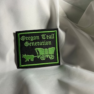 May include: A black and green embroidered patch with the text "Oregon Trail Generation" and a green pixelated image of a covered wagon pulled by an ox.