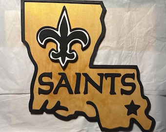 May include: A wooden cutout of the state of Louisiana with the New Orleans Saints logo and the word "SAINTS" in black lettering. The state is painted gold with a black outline.