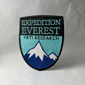 May include: A black and blue embroidered patch with the text "Expedition Everest Yeti Research" and a stylized mountain range with snow-capped peaks.