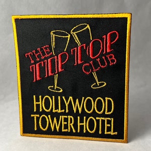 May include: A black embroidered patch with a gold border. The patch features a red embroidered design of two champagne glasses with the text "The Top Top Club" and "Hollywood Tower Hotel" in gold.