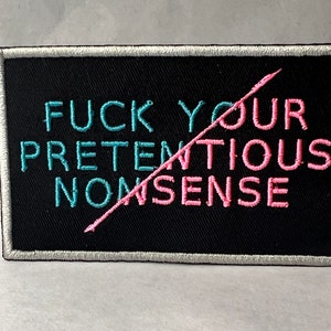 May include: Embroidered patch with the phrase "FUCK YOUR PRETENTIOUS NONSENSE" in teal and pink lettering on a black background. The words are crossed out with a pink line. The patch has a silver border.