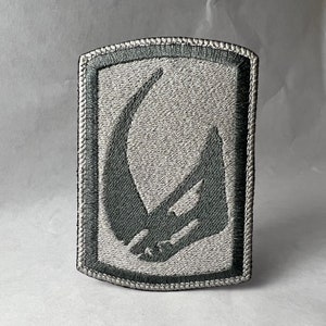 May include: A grey embroidered patch featuring a dark grey Mandalorian symbol. The patch has a white border and is stitched with grey thread.