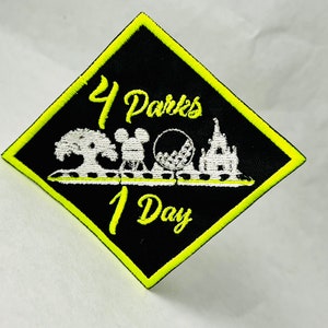 May include: A black diamond-shaped patch with a yellow border. The patch features white embroidered text that reads "4 Parks 1 Day" and a white embroidered image of a park with a tree, a castle, a Ferris wheel, and a Mickey Mouse head.