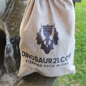 May include: A beige burlap drawstring bag with a black dinosaur skull logo and the text "DINOSAUR21.COM STEPPING BACK IN TIME." The bag is sitting on a wet, weathered surface, with a blurred background of water and greenery.