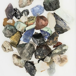 Pode incluir: Uma cole&ccedil;&atilde;o de v&aacute;rias pedras preciosas brutas e n&atilde;o polidas em uma variedade de cores, incluindo verde, azul, roxo, vermelho e marrom. As pedras est&atilde;o dispostas em uma pilha sobre um fundo branco.