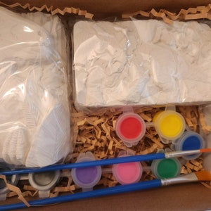 May include: A craft kit with two white plaster molds, six small paint pots in red, yellow, blue, green, purple, and pink, and two paintbrushes. The kit is packaged in a brown cardboard box with shredded paper.