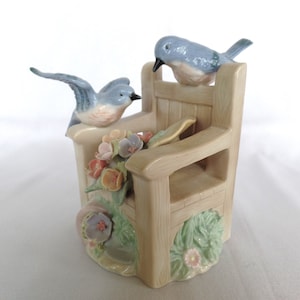 May include: A porcelain figurine of a wooden chair with two bluebirds perched on the back and arms. The chair is surrounded by flowers and greenery.