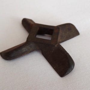 May include: A rusty, metal meat grinder blade with a square center and four blades extending outward. The blade is a dark brown color and appears to be vintage. The object is set against a white background.