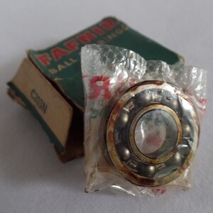 May include: A metallic ball bearing with visible steel spheres inside a circular housing, enclosed in a clear plastic wrapper. Behind it sits a green and tan cardboard box labeled FAFNIR BEARINGS with the model number C203N printed on the side.