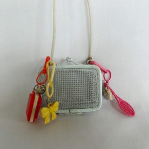 May include: A vintage-style necklace featuring a small, light blue mesh purse pendant. The necklace includes various charms: a red and white rectangle, a yellow bow, a silver bell, and a pink tennis racket. The necklace is suspended from a white cord.