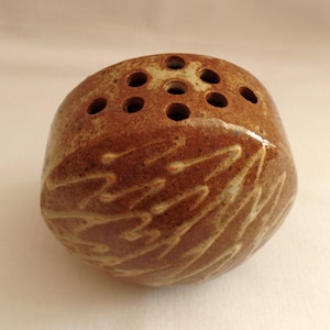 May include: A brown ceramic flower frog with multiple holes on top for arranging stems. The rounded form features a textured surface with a leaf-like pattern carved into the side. A unique piece for floral arrangements.