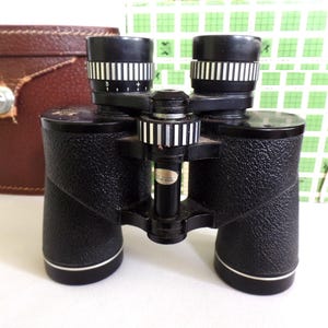 May include: Black binoculars with a leather case. The binoculars have a silver trim and a focusing wheel on each lens.