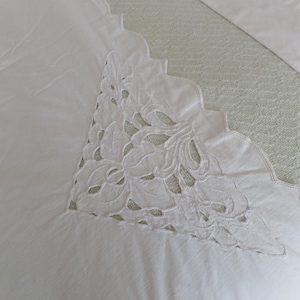 May include: Close-up of a white, embroidered textile with a scalloped edge. The embroidery features a floral design with cut-out details. The fabric appears to be a soft cotton or linen, suitable for bedding or decorative use.