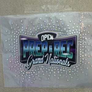 May include: A clear, rectangular sticker with the text "OPEN PREP & REC Grand Nationals" in a teal and purple design. The sticker is embellished with numerous iridescent rhinestones, creating a sparkling effect. The background is a neutral, textured surface.