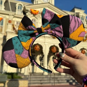 May include: A pair of black, purple, yellow, and teal patchwork mouse ears with a large bow. The ears are made of fabric and have a stitched design.
