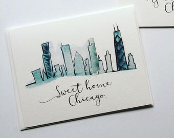 May include: A watercolor painting of the Chicago skyline with the text "Sweet home Chicago." The painting features a blue and green color scheme and depicts several buildings, including the Willis Tower.