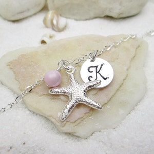 Puede incluir: Un brazalete de cadena de plata con una cuenta rosa, un dije de estrella de mar de plata y un dije de disco de plata con la letra "K" grabada.