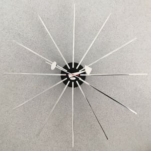 Op de afbeelding: Een moderne wandklok met een zwart cirkelvormig midden en zilverkleurige stralende armen. De wijzers zijn wit en rood, en de wijzerplaat heeft een minimalistisch ontwerp. De armen van de klok zijn ongeveer 30 cm lang.