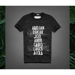 May include: Dark gray t-shirt with the names of African cities in white block letters: Abidjan, Dakar, Jozi, Addis, Cairo, Lagos, and Accra. The shirt has a crew neck and short sleeves.