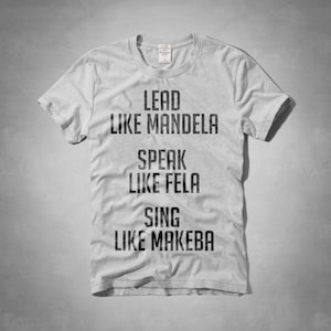 Peut inclure: T-shirt gris clair avec l'inscription noire : « LEAD LIKE MANDELA, SPEAK LIKE FELA, SING LIKE MAKEBA ». Le t-shirt a des manches courtes et un col rond.