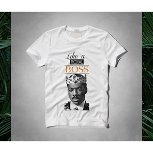May include: White t-shirt with the text "Like a ROYAL BOSS" in black and orange. The shirt features a black and white portrait of a man wearing a crown. The t-shirt is a crew neck style.