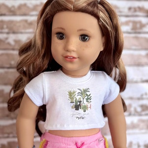 May include: A white crop top with short sleeves features a graphic of potted plants and the text "Just one more". The doll is wearing pink sweatpants with a rainbow stripe down the side.