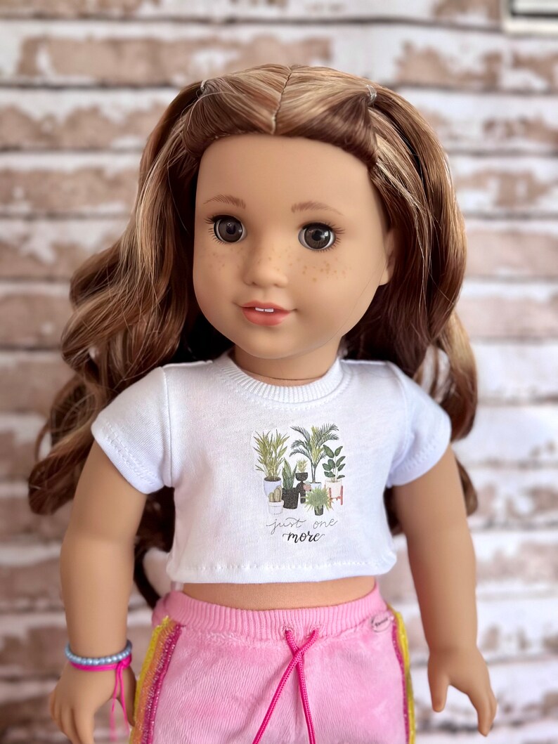 May include: A white crop top with short sleeves features a graphic of potted plants and the text "Just one more". The doll is wearing pink sweatpants with a rainbow stripe down the side.