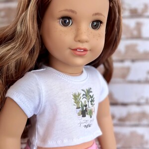 May include: A doll wearing a white t-shirt with a graphic of potted plants and the text "just grow more". The doll is also wearing pink shorts with a rainbow stripe.
