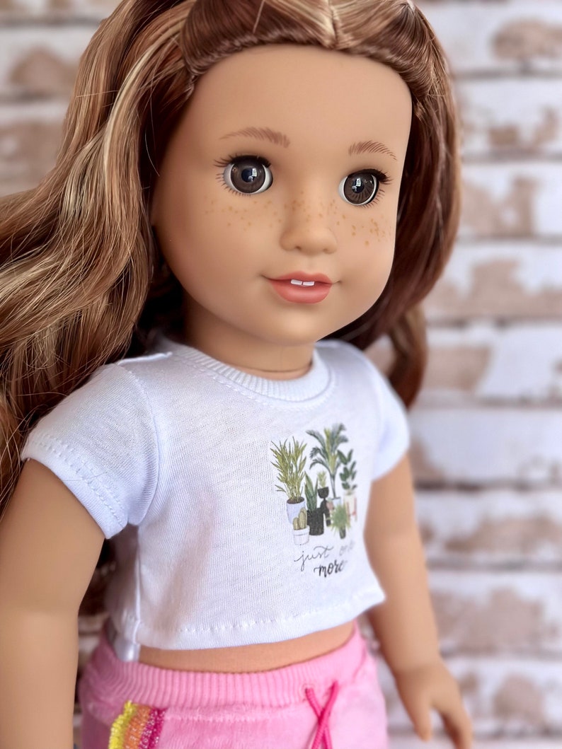 May include: A doll wearing a white t-shirt with a graphic of potted plants and the text "just grow more". The doll is also wearing pink shorts with a rainbow stripe.