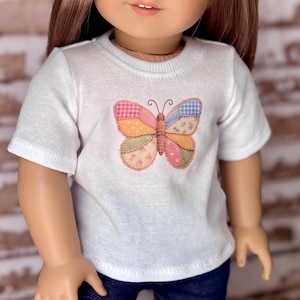 May include: A white short-sleeved t-shirt featuring a central butterfly graphic. The butterfly is composed of various patchwork-style segments in shades of pink, orange, blue, and green, with a brown body and two thin antennae at the top center.