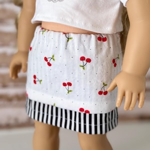 May include: A doll wearing a white crop top with a cherry graphic and a white skirt. The skirt features a cherry print with black polka dots and a black and white striped hem. The doll is wearing a matching outfit.