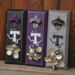 May include: Three wooden bottle openers in black, purple, and gray. Each has a metal opener and a Texas-shaped logo with a "T" inside. Bottle caps are attached below each opener. The openers are mounted on a wooden surface.