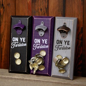 May include: Three wooden bottle openers with a magnetic back to hold bottle caps. Each opener has a different color: black, purple, and gray. The openers have the text "On Ye Tarleton" printed on them.