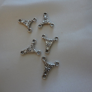 May include: Five silver-colored, ornate connector charms. Each charm has a triangular shape with three circular holes and decorative floral patterns. The charms are scattered on a light-colored, slightly textured surface.