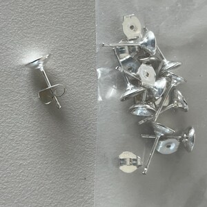 May include: A collection of silver-colored earring posts with butterfly backs. The posts have a flat, round top and a slender post. The earring backs are butterfly-shaped. The earrings are displayed on a light gray surface.