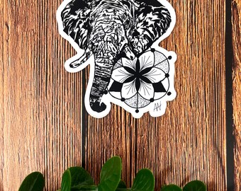 May include: Black and white sticker of an elephant's head with a geometric mandala design. The elephant is facing left and has a detailed, textured design. The mandala is in the shape of a flower with a dot pattern and a black triangle in the center.