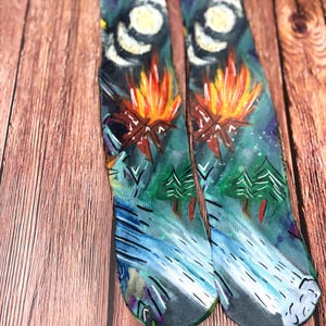 May include: A pair of socks with a colorful, artistic design. The socks feature a campfire scene with a starry night sky, a full moon, and a flowing river. The color palette includes blues, greens, oranges, and white.