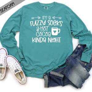 May include: A teal long-sleeved t-shirt with the text "It's a Fuzzy Socks & Hot Cocoa Kinda Night" in white. The shirt is shown on a white background with a pair of white sneakers and blue jeans.
