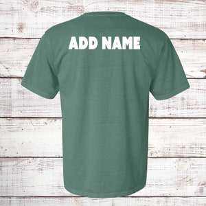 May include: A short-sleeved, teal-colored t-shirt with the text "ADD NAME" printed in white across the upper back. The shirt is made of a soft material and is displayed against a white wooden background.