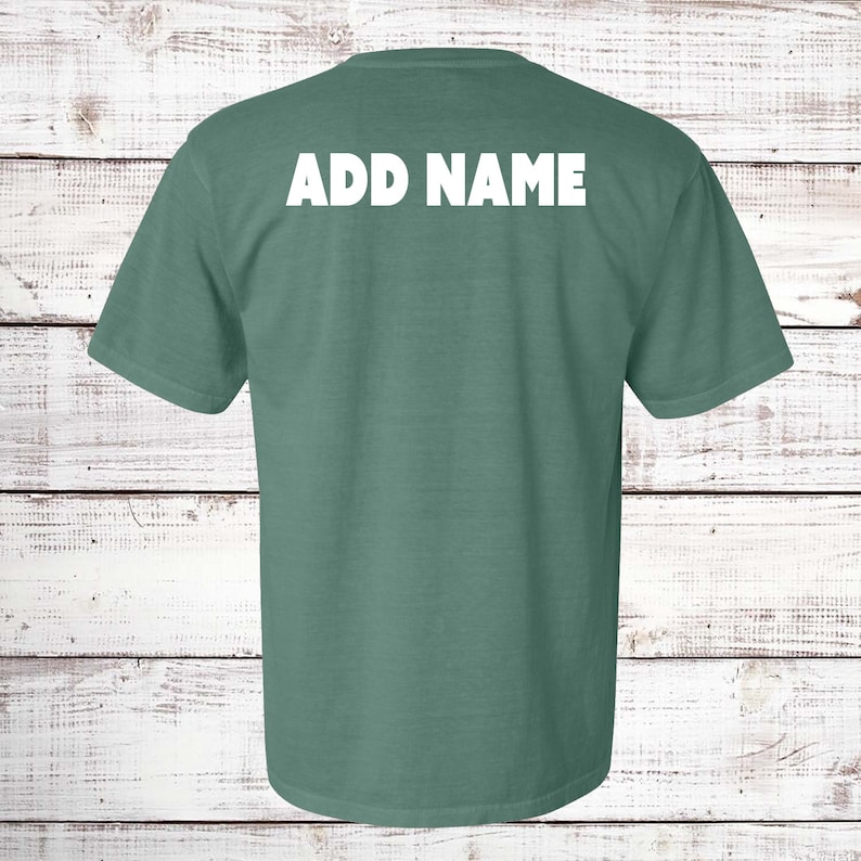 May include: A short-sleeved, teal-colored t-shirt with the text "ADD NAME" printed in white across the upper back. The shirt is made of a soft material and is displayed against a white wooden background.