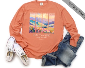 May include: Terracotta long-sleeve shirt with a landscape graphic and the text "TRUST in the LORD with all your heart". The shirt is paired with blue jeans and white canvas sneakers.