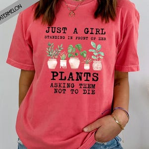 May include: A red t-shirt with a graphic of potted plants and the text "Just a girl standing in front of her plants asking them not to die."