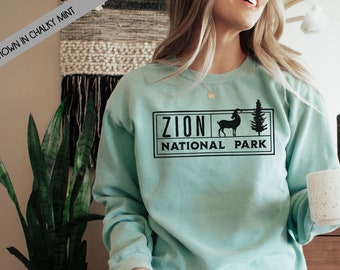 May include: A light blue sweatshirt with a black graphic design that says "Zion National Park" with a silhouette of a mountain goat and a pine tree.