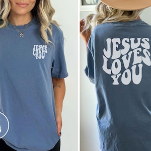 May include: A blue jean colored short-sleeve t-shirt featuring the white text Jesus Loves You printed in a wavy font on the left chest and in a larger size across the back. The shirt has a crew neckline and a relaxed fit.