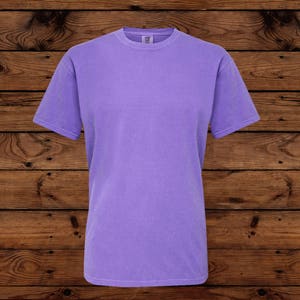 May include: A solid lavender short-sleeve t-shirt. The shirt has a crew neck and a relaxed fit. The fabric appears soft and slightly faded. The background is a dark wood grain.