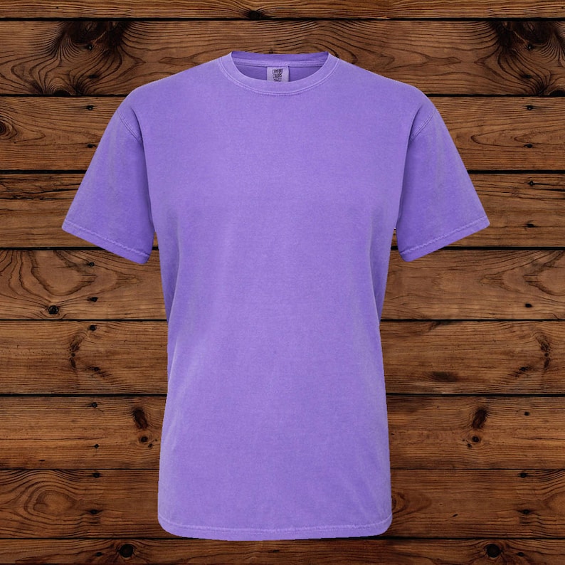 May include: A solid lavender short-sleeve t-shirt. The shirt has a crew neck and a relaxed fit. The fabric appears soft and slightly faded. The background is a dark wood grain.